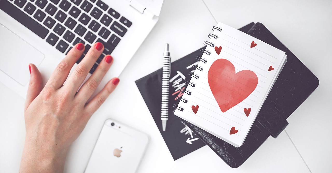 woman hand red painted nails on keyboard with drawn hearth on paper by side